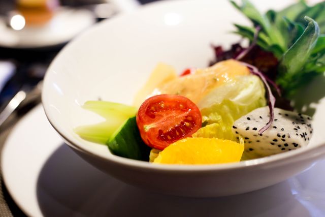 This image captures a fresh mixed salad served in a white bowl featuring vibrant vegetables and fruits such as cherry tomatoes, cucumber, lettuce, oranges, and greens. Ideal for use in articles, advertisements, and blogs focused on healthy eating, nutrition, meal planning, vegan, vegetarian, and health food promotions. Also suitable for culinary publications, recipe books, and restaurant menus advertising nutritious meal options.