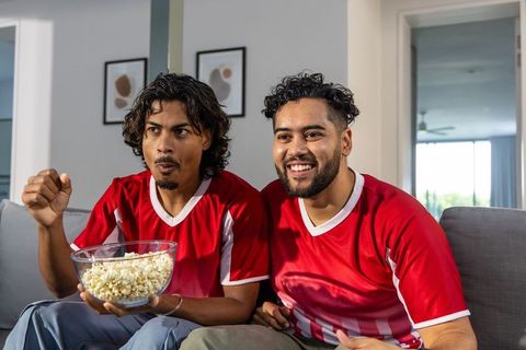 Friends watching soccer match at home wearing red jerseys eating popcorn and cheering