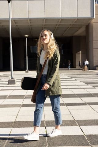 Young woman walking with skateboard sleeve and headphones in modern urban plaza