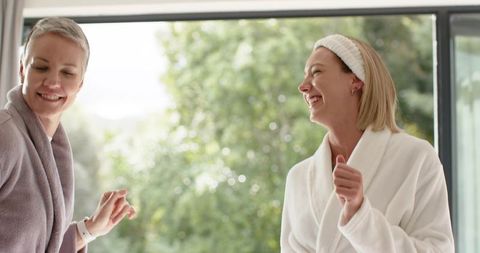 Two women laughing in bathrobes by sliding glass door with smartwatch and morning greenery