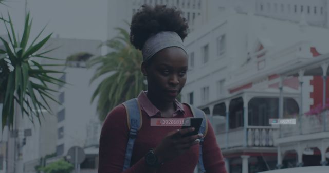 Young African woman engaged with her smartphone while walking down a vibrant city street. She wears casual clothing and carries a backpack, suggesting she is a student or commuter. Use this image for themes involving urban life, technology, millennials, or outdoor communication.