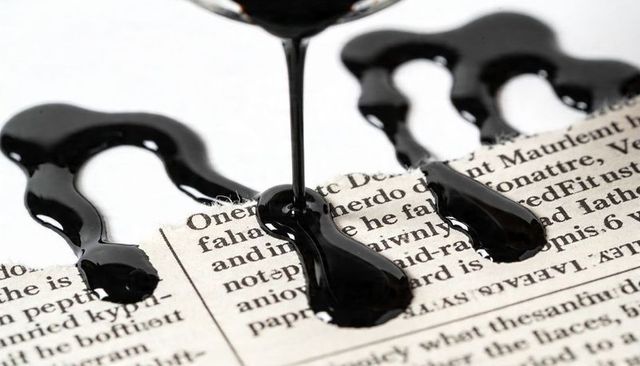 Glossy black viscous liquid pouring on newspaper forming shiny puddles