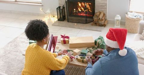 Black couple wrapping christmas gifts by fireplace, cozy living room ribbon and present prep