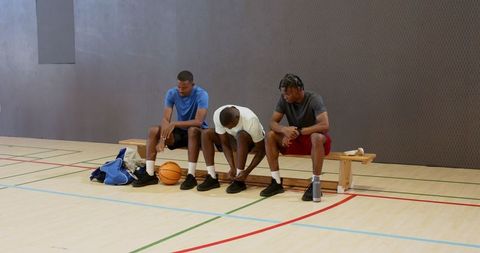 Three teammates resting on gym bench tying shoes with basketball on court