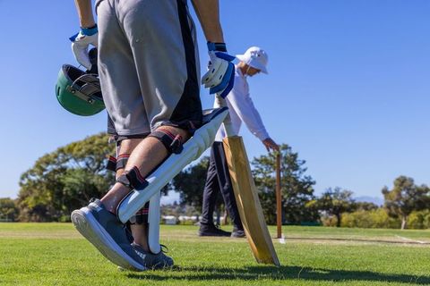 Close-up batter standing in pads holding helmet and bat near stumps on sunny cricket field