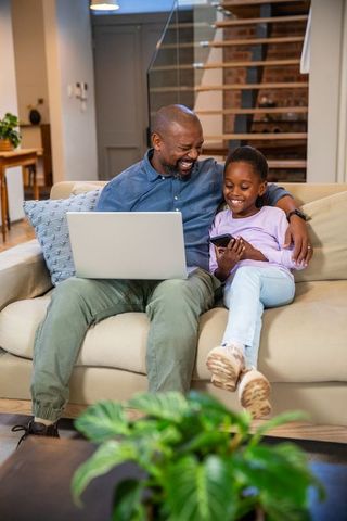 African american father and daughter sharing phone and laptop on couch, laughing together