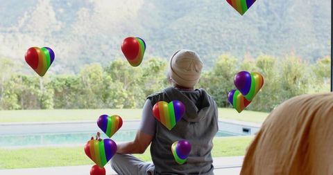 Sitting asian man wearing beanie and hoodie facing pool patio with rainbow heart balloons