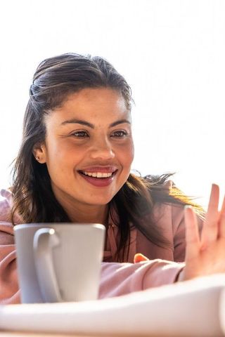 Woman in 20s smiling and reaching toward camera with mug in cozy backlit morning