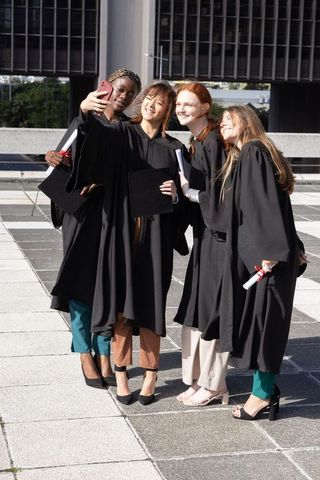 Diverse graduates taking selfie on urban campus plaza wearing gowns holding diplomas