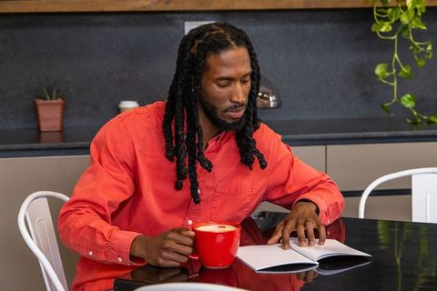 African american man reading book while holding red coffee mug at modern kitchen table
