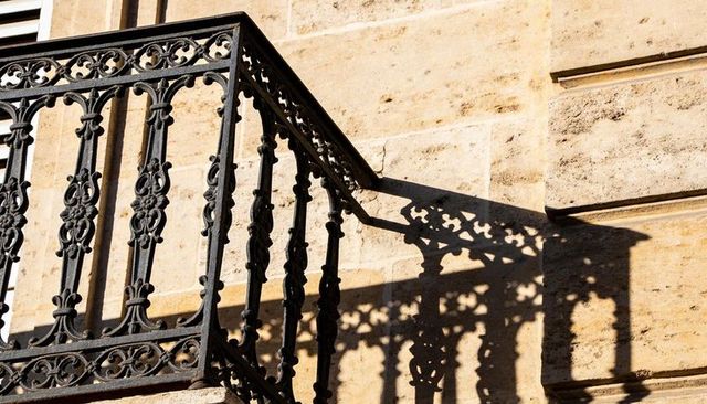 Sunlit ornamental wrought iron balcony casting intricate shadow on limestone facade detail