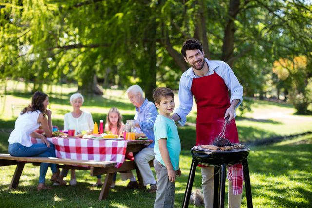 Father and son enjoying a barbecue in a park with family members sitting at a picnic table in the background. Ideal for promoting family activities, outdoor events, summer gatherings, and leisure time. Perfect for advertisements, brochures, and websites focused on family bonding, outdoor fun, and healthy living.