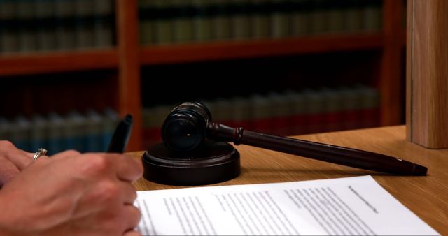 A Caucasian middle-aged individual, a judge or lawyer, is signing a document, with a gavel in the foreground symbolizing legal authority. The presence of law books in the background suggests a formal legal setting, emphasizing the importance of the documentation being handled.