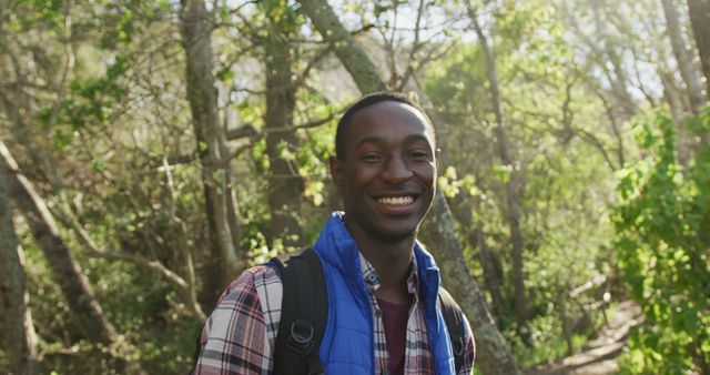 Young African American male enjoying hiking in lush forest with warm sunlight filtering through trees. Casual plaid shirt and blue vest highlight relaxed and adventurous spirit. Perfect for illustrating outdoor activities, nature travel, leisure time, and active lifestyles.