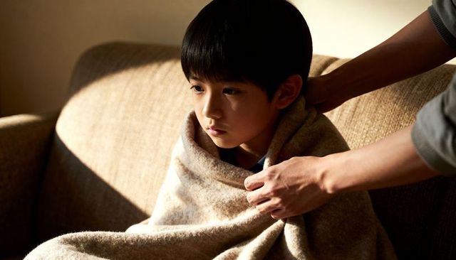 Wrapping asian child in cozy blanket, caregiver comforting on sunlit sofa