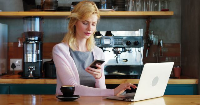 Blonde woman working in coffee shop typing on laptop and checking phone, looks focused and professional, ideal for illustrating concepts of remote work, modern technology, or business productivity in a casual urban setting.