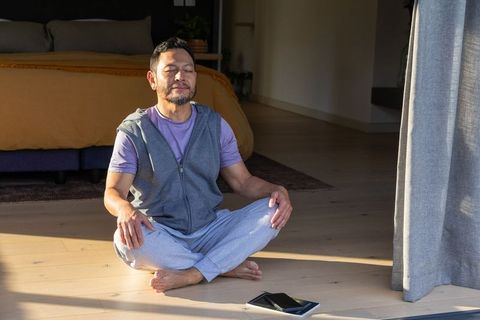 Mature man meditating in sunlit minimalist bedroom, relaxed morning routine with tablet
