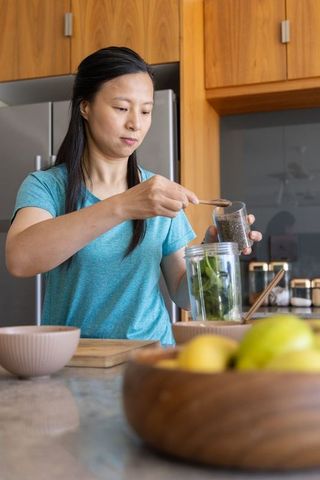 Asian woman adding chia seeds to spinach smoothie in modern wooden kitchen with fruit bowl