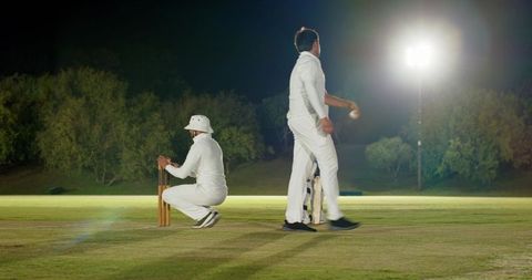 Night cricket under floodlights with bowler delivering and wicketkeeper crouching on outfield