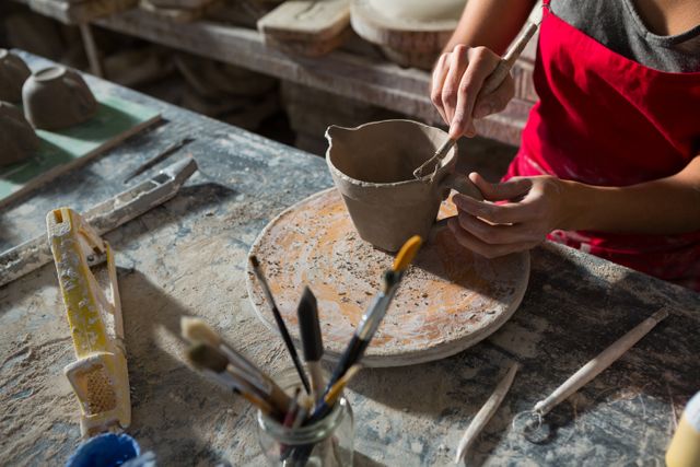 Female potter carving a clay mug in a studio, showcasing detailed craftsmanship. Ideal for use in articles about pottery, artisan crafts, creative hobbies, or handmade goods. Can be used to illustrate the process of ceramic art or the dedication of artists to their craft.
