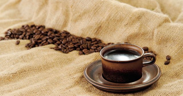 A steaming cup of coffee sits beside a trail of coffee beans on a burlap surface, with copy space. The arrangement suggests a rustic and warm atmosphere, ideal for coffee enthusiasts and morning rituals.