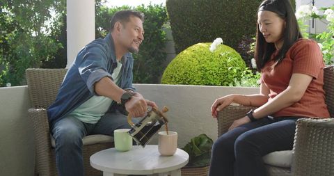 Asian couple pouring coffee on sunlit patio, enjoying relaxed morning conversation