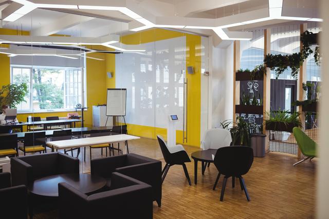 This image showcases a modern office interior featuring bright yellow walls and contemporary furniture. The space includes various seating options such as sofas, chairs, and tables, along with indoor plants that add a touch of greenery. The large windows allow natural light to flood the room, creating a bright and inviting atmosphere. This image can be used for articles or advertisements related to office design, modern workspaces, business environments, or professional settings.