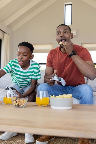 African american father and son playing video games sharing snacks on living room couch