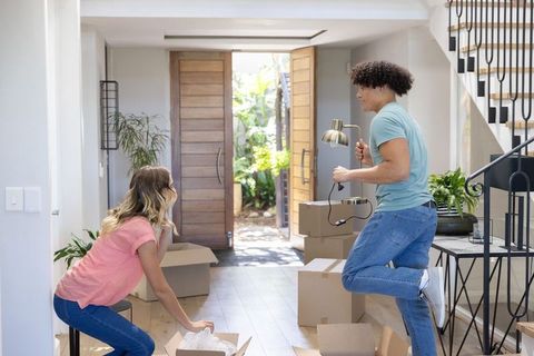 Multiracial couple unpacking boxes in sunlit foyer, carrying lamp and arranging new home