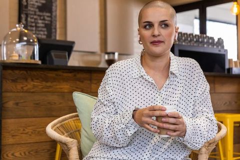 Bald woman sitting in cozy cafe holding takeaway cup wearing polka-dot blouse