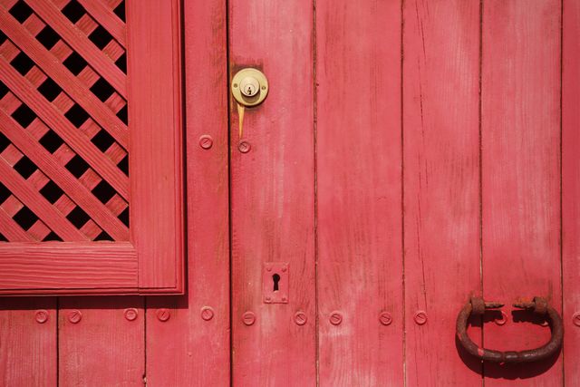 This image depicts a vintage red wooden door with a rusted handle, lock, keyhole, and lattice window panel. Ideal for themes related to architecture, history, antique decor, rustic aesthetics, and weathering. Can be used in blog posts about vintage homes, DIY restoration projects, and historical architecture, or as a background in design projects seeking a rustic feel.