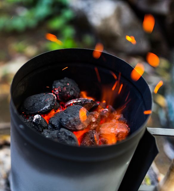 Glowing charcoal in a chimney starter with sparks flying. Ideal for conveying themes of outdoor cooking, grilling, barbecuing, or campfire preparations. Great for use in cooking tutorials, outdoor activity promotions, camping ads, or in food and cooking blogs.