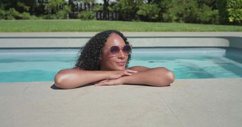 African american woman relaxing at backyard pool wearing sunglasses and swimwear, sunlit leisure