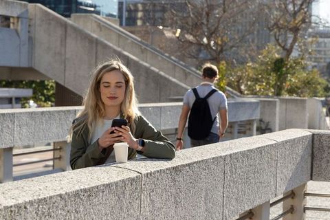 Woman checking smartphone while leaning on concrete railing with coffee, man walking in urban plaza