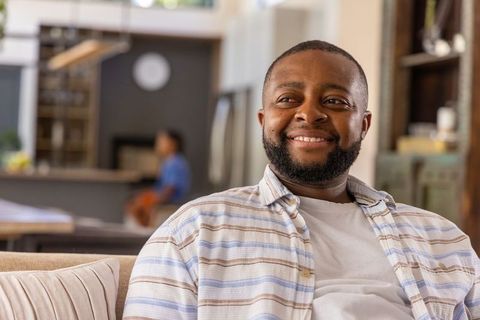 African american man smiling on couch in cozy open-plan living room with kitchen background