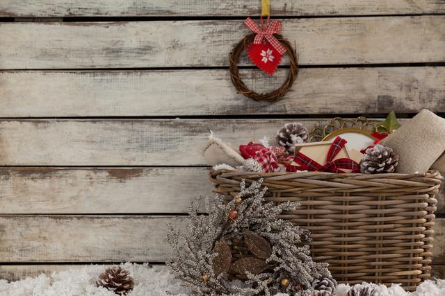 Wooden backdrop with cozy Christmas setting featuring a wicker basket filled with holiday decorations such as pinecones, wrapped gifts, and ornaments. A small wreath with a heart ornament hangs on the wooden wall. Ideal for holiday greeting cards, festive advertisements, and home decor inspiration.
