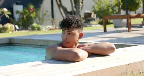 Young man relaxing at sunlit backyard pool edge on wooden deck, garden leisure