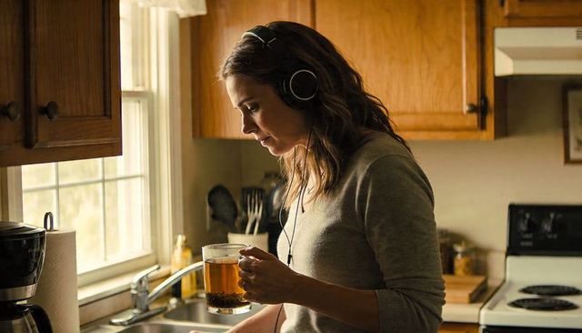Woman holding glass mug of tea listening to headphones by sunlit kitchen sink