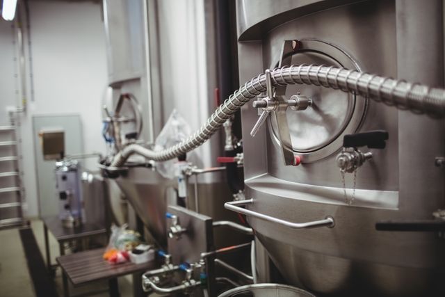Image shows industrial brewery equipment with a focus on a metallic pipe connected to a large stainless steel tank. Ideal for use in articles or advertisements related to beer production, brewery processes, or industrial manufacturing. Can be used to illustrate modern brewing technology and equipment in commercial breweries.