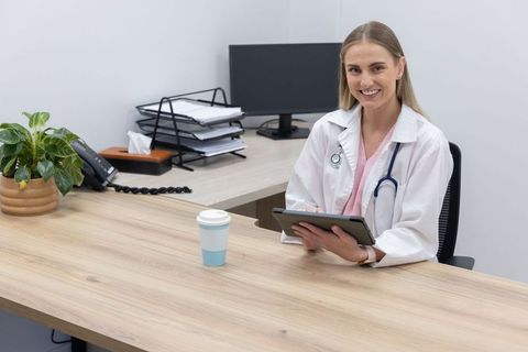 Smiling female physician holding tablet at clinic desk with stethoscope, coffee and plant