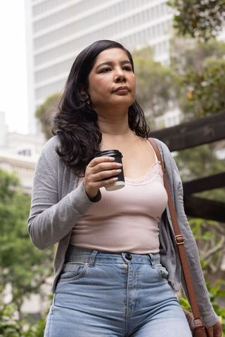 Woman walking through downtown plaza holding takeout coffee cup, casual outfit, urban commute