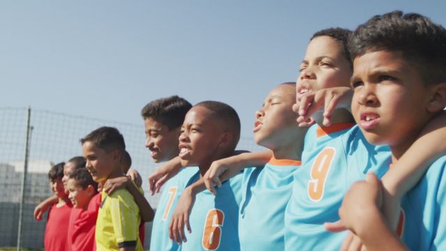 The video shows a group of young, diverse male soccer players wearing team uniforms, standing on a soccer field with arms around each other, singing joyfully. Ideal for promoting youth sports programs, unity and diversity in teams, community projects, and advertisements that emphasize teamwork and sportsmanship.