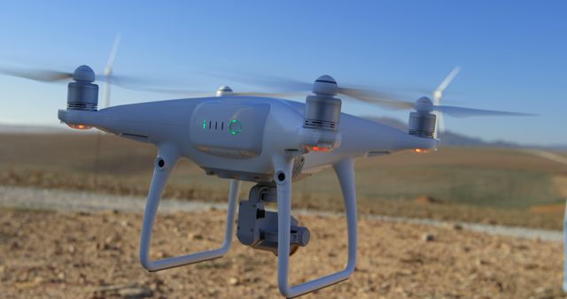 A drone equipped with a camera hovering in a rural setting with wind turbines in the background. Useful for content related to technology advancements, aerial photography, renewable energy, eco-friendly innovations, and outdoor exploration.