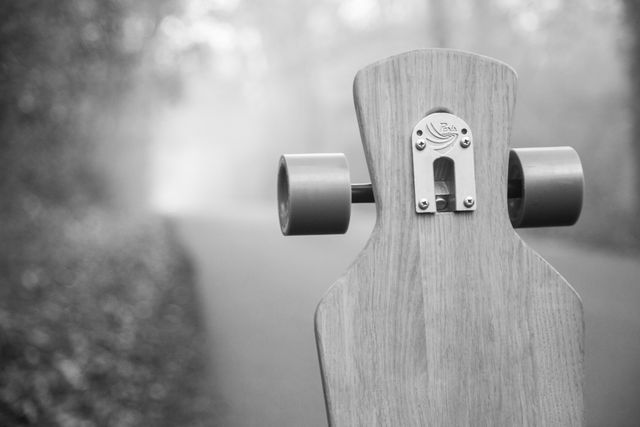 Close-up of a wooden longboard with wheels, positioned on an asphalt path in a misty, foggy forest. Suitable for illustrating themes of adventure, outdoor sports, skateboarding, and the beauty of nature. Perfect for promoting skateboarding gear, nature exploration, or wood craftwork.