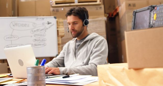 Warehouse worker is using laptop with headset, focusing on managing inventory. Ideal for visualizations of supply chain management, warehouse technology, organizational tasks, and remote communication in logistics businesses.