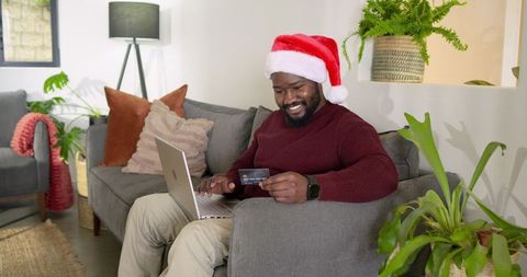 African american man wearing santa hat doing online holiday shopping on laptop at home