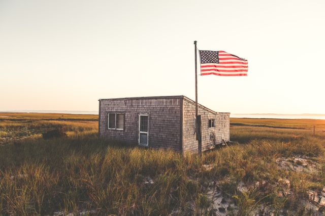 Rustic cabin situated in wide open field with American flag waving. Ideal for scenes depicting rural life, patriotic themes, and American countryside. Perfect for travel blogs, patriotism articles, and nature magazines.