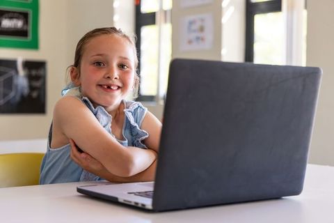 Smiling schoolgirl using laptop in bright classroom for online learning and digital study
