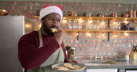 African American man wearing Santa hat eating homemade Christmas cookies in cozy kitchen