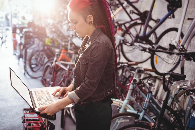 Young female mechanic with red hair using a laptop in a bicycle repair shop. Surrounding her are various bikes, indicating a busy workspace. Useful for themes involving technology, mechanical work, female workers in traditionally male-dominated fields, and modern repair shops.